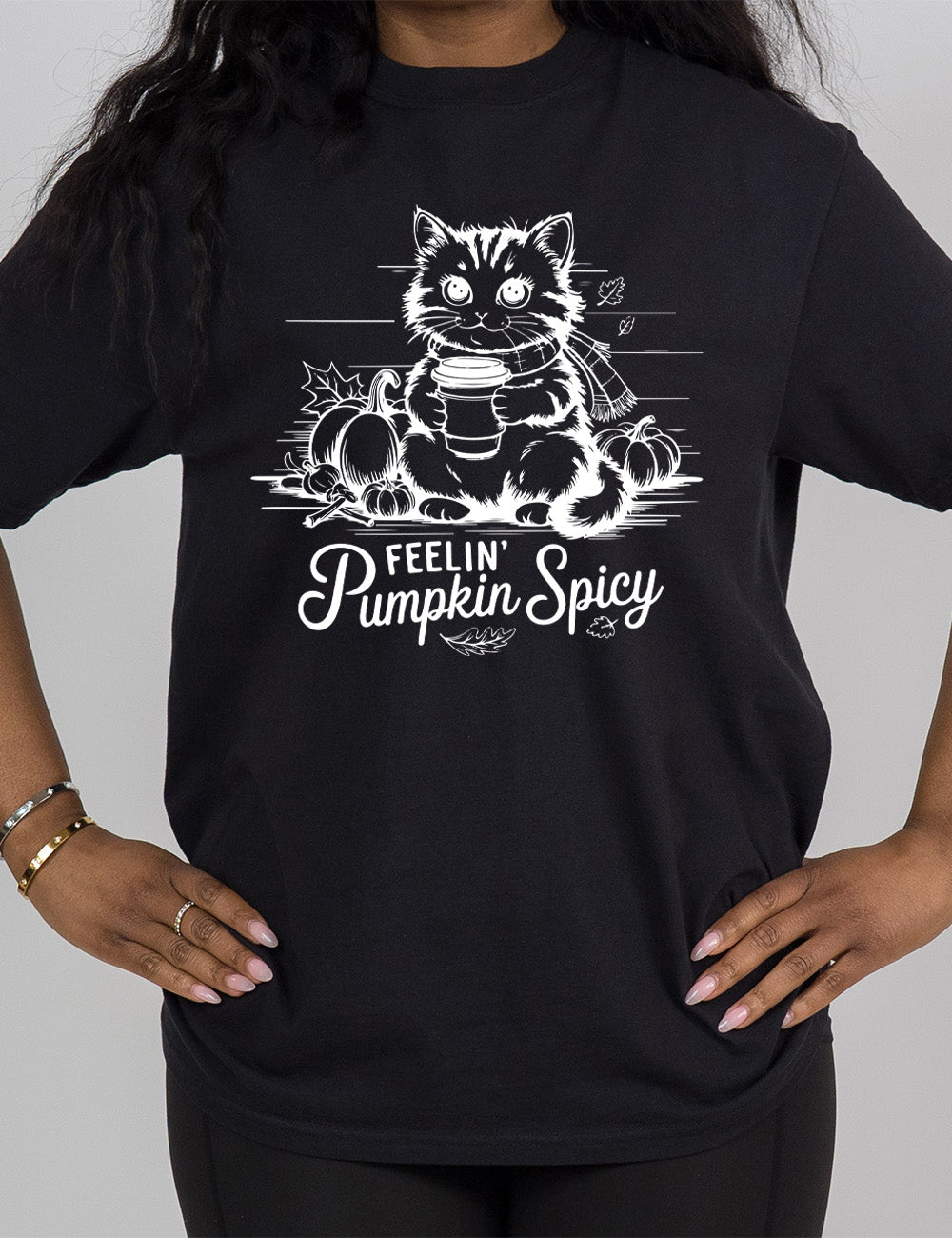 black modern tee with the graphic of a cat, wearing a scarf, next to a few pumpkins of various sizes and a few falling leaves, holding a takeaway coffee, and the words underneath saying "feelin' pumpkin spicy"