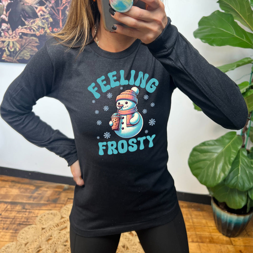 heather black long sleeve shirt with the words "feeling frosty" on it, with the graphic of snowperson, wearing a hat and scarf, drinking an iced drink, surrounded by snowflakes