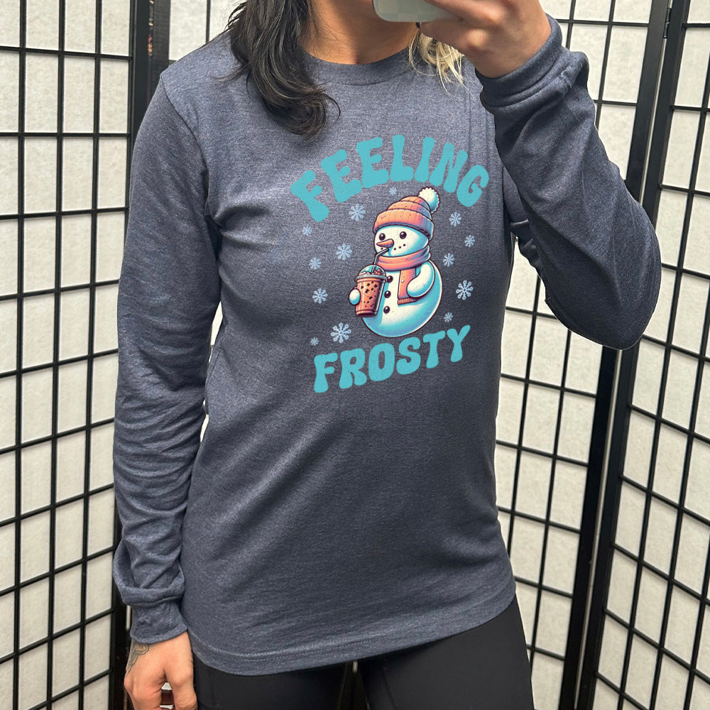 heather navy long sleeve shirt with the words "feeling frosty" on it, with the graphic of snowperson, wearing a hat and scarf, drinking an iced drink, surrounded by snowflakes