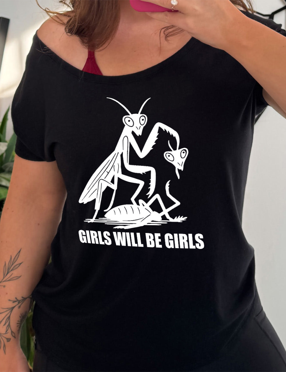 black slouchy tee with the graphic of a praying mantis, holding up the head of the other praying mantis, whose body she is stepping on, with the words underneath it saying "girls will be girls"