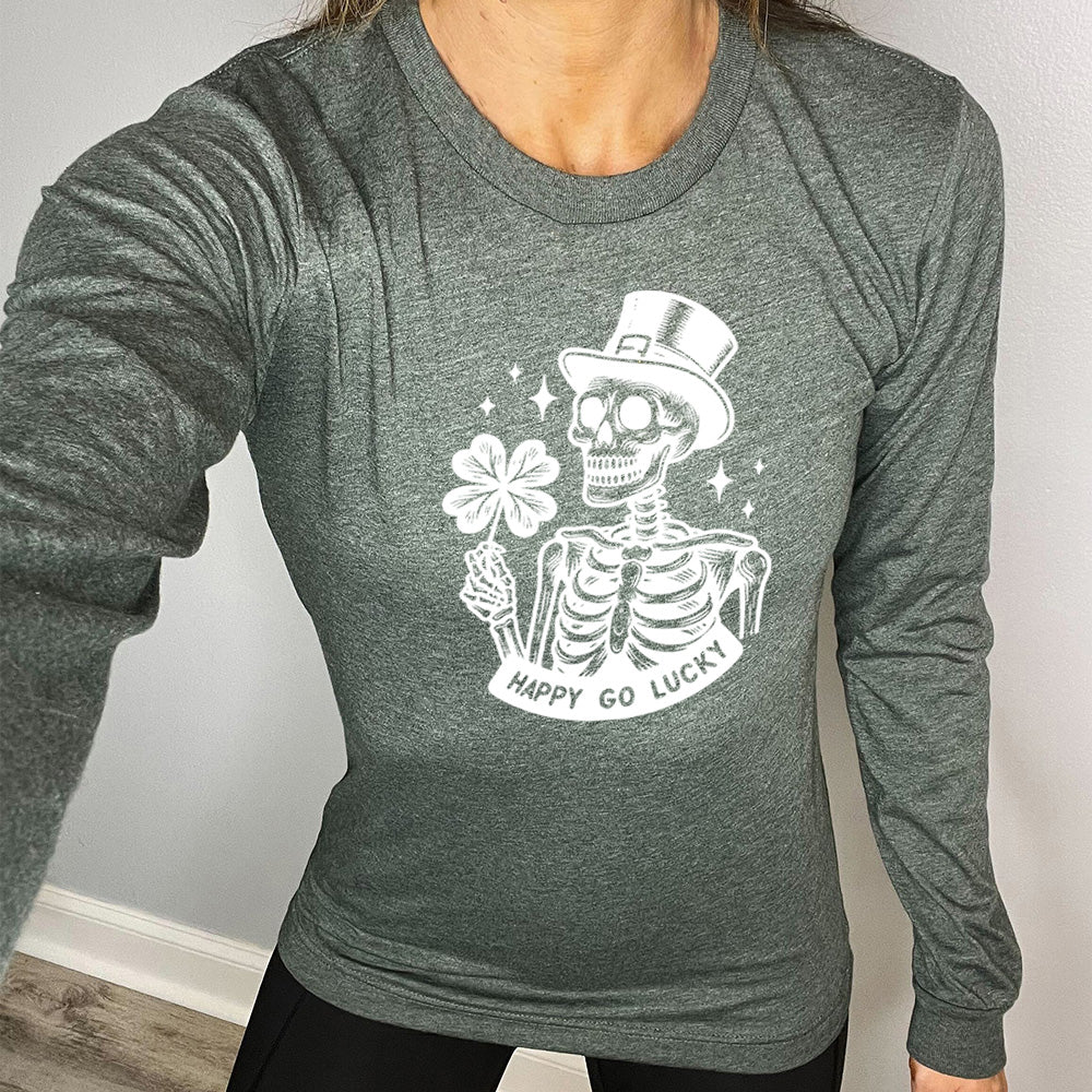 heather forest long sleeve shirt with the graphic of a skeleton wearing a leprechaun hat and holding a four-leaf clover, with a few little stars around, and the text "happy go lucky" underneath