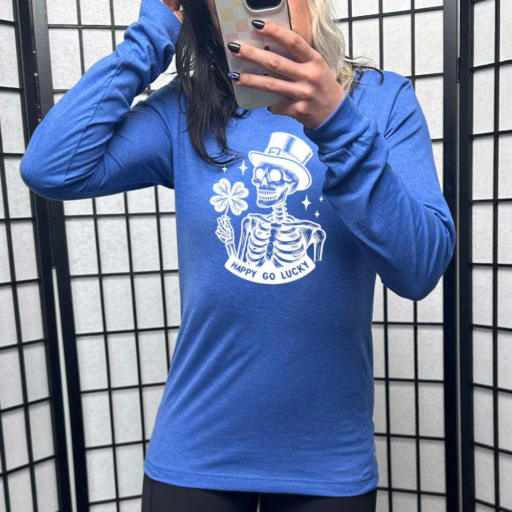 heather true royal long sleeve shirt with the graphic of a skeleton wearing a leprechaun hat and holding a four-leaf clover, with a few little stars around, and the text "happy go lucky" underneath