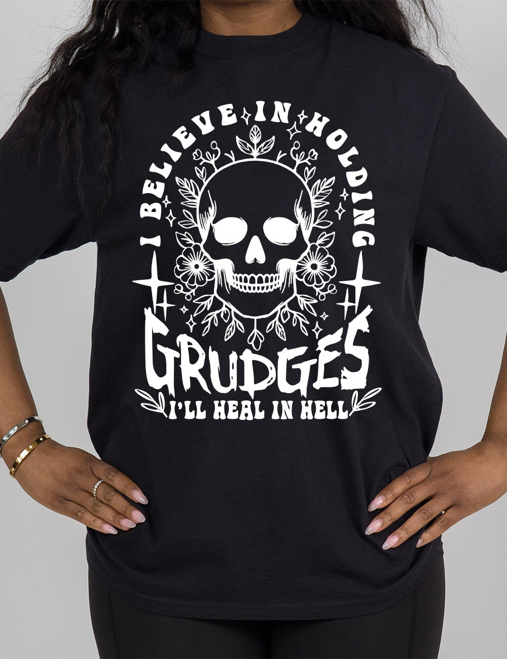 black modern tee with print placement on the front, with the graphic of a skull among flowers, and words arching over it and words underneath, saying "I believe in holding GRUDGES I'll heal in hell"