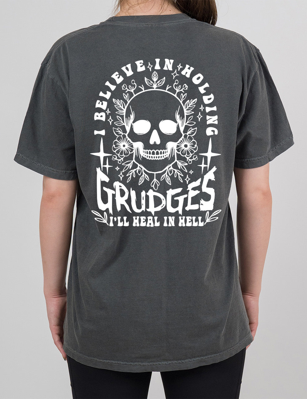 pepper modern tee with print placement on the back, with the graphic of a skull among flowers, and words arching over it and words underneath, saying "I believe in holding GRUDGES I'll heal in hell"