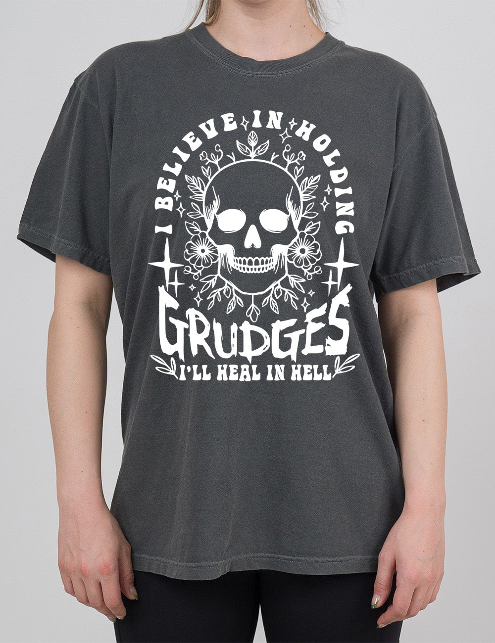 pepper modern tee with print placement on the front, with the graphic of a skull among flowers, and words arching over it and words underneath, saying "I believe in holding GRUDGES I'll heal in hell"