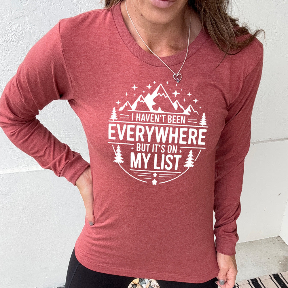 heather clay long sleeve with the words "I haven't been everywhere but it's on my list" as part of a circular design involving the graphic of small mountains on top and a few trees on the sides and stars scattered around