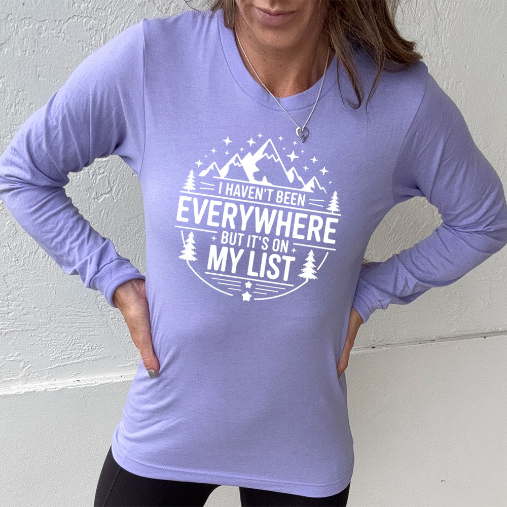 heather dark lavender long sleeve with the words "I haven't been everywhere but it's on my list" as part of a circular design involving the graphic of small mountains on top and a few trees on the sides and stars scattered around