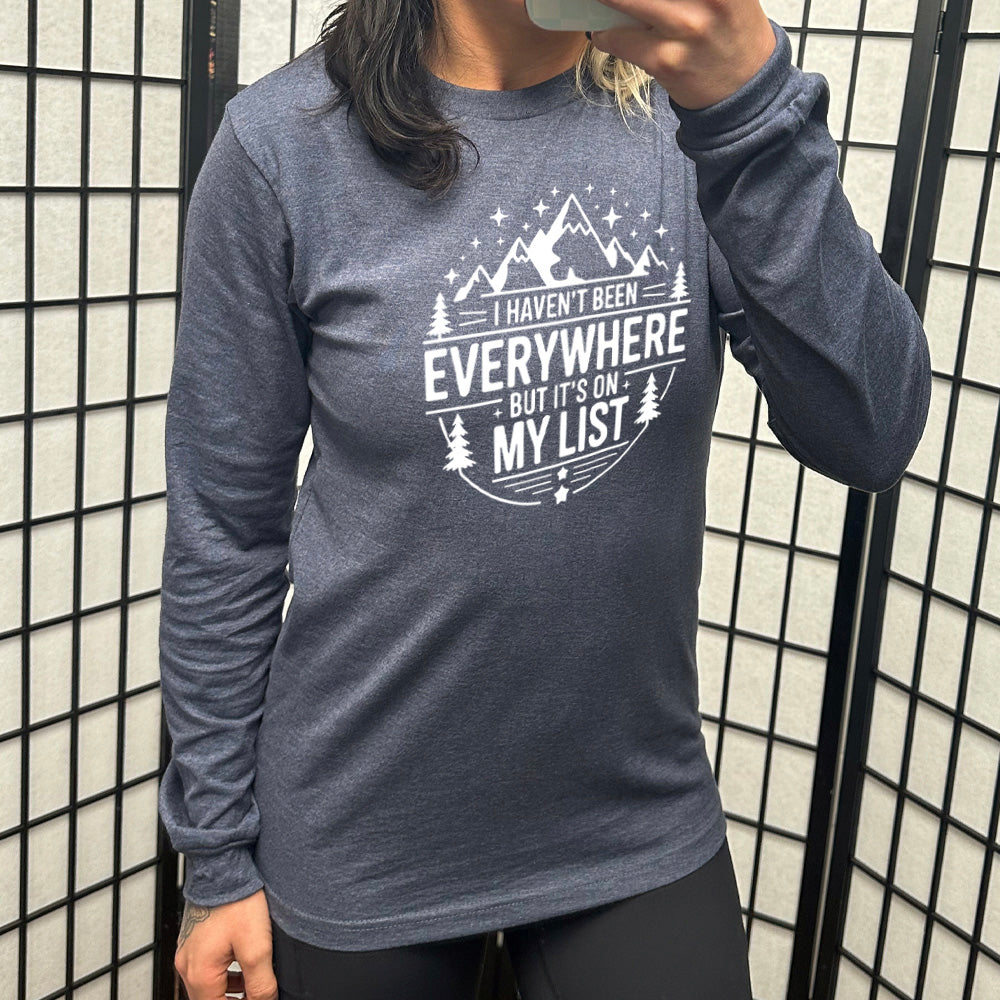 heather navy long sleeve with the words "I haven't been everywhere but it's on my list" as part of a circular design involving the graphic of small mountains on top and a few trees on the sides and stars scattered around