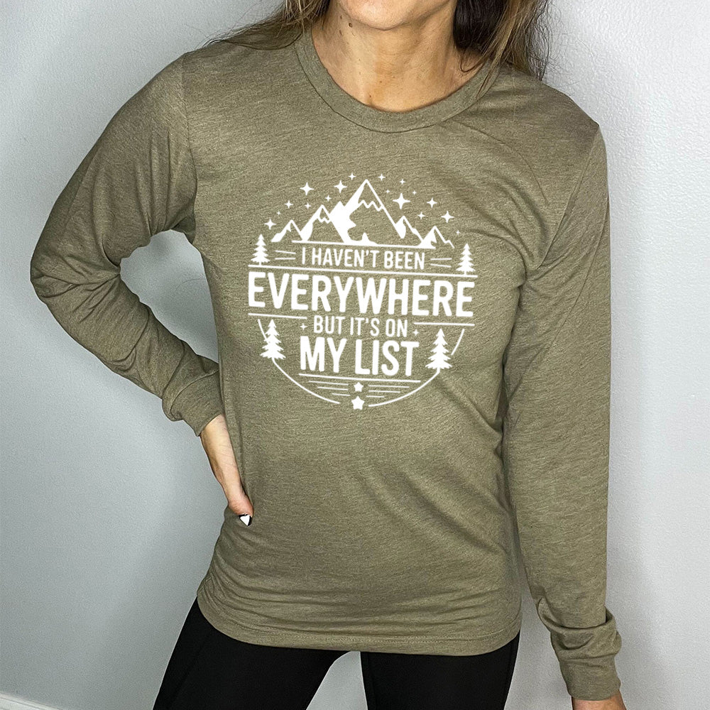heather olive long sleeve with the words "I haven't been everywhere but it's on my list" as part of a circular design involving the graphic of small mountains on top and a few trees on the sides and stars scattered around