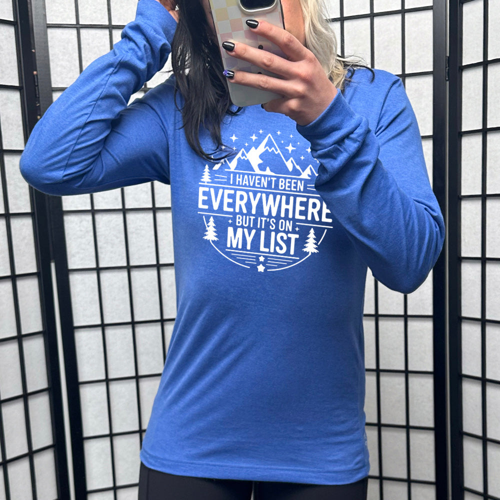 heather true royal long sleeve with the words "I haven't been everywhere but it's on my list" as part of a circular design involving the graphic of small mountains on top and a few trees on the sides and stars scattered around