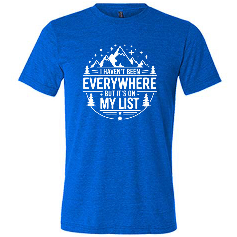blue unisex tee with the words "I haven't been everywhere but it's on my list" as part of a circular design involving the graphic of small mountains on top and a few trees on the sides and stars scattered around