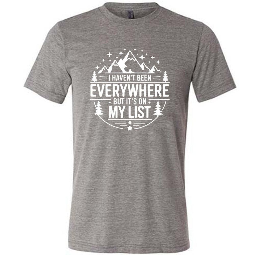 grey unisex tee with the words "I haven't been everywhere but it's on my list" as part of a circular design involving the graphic of small mountains on top and a few trees on the sides and stars scattered around