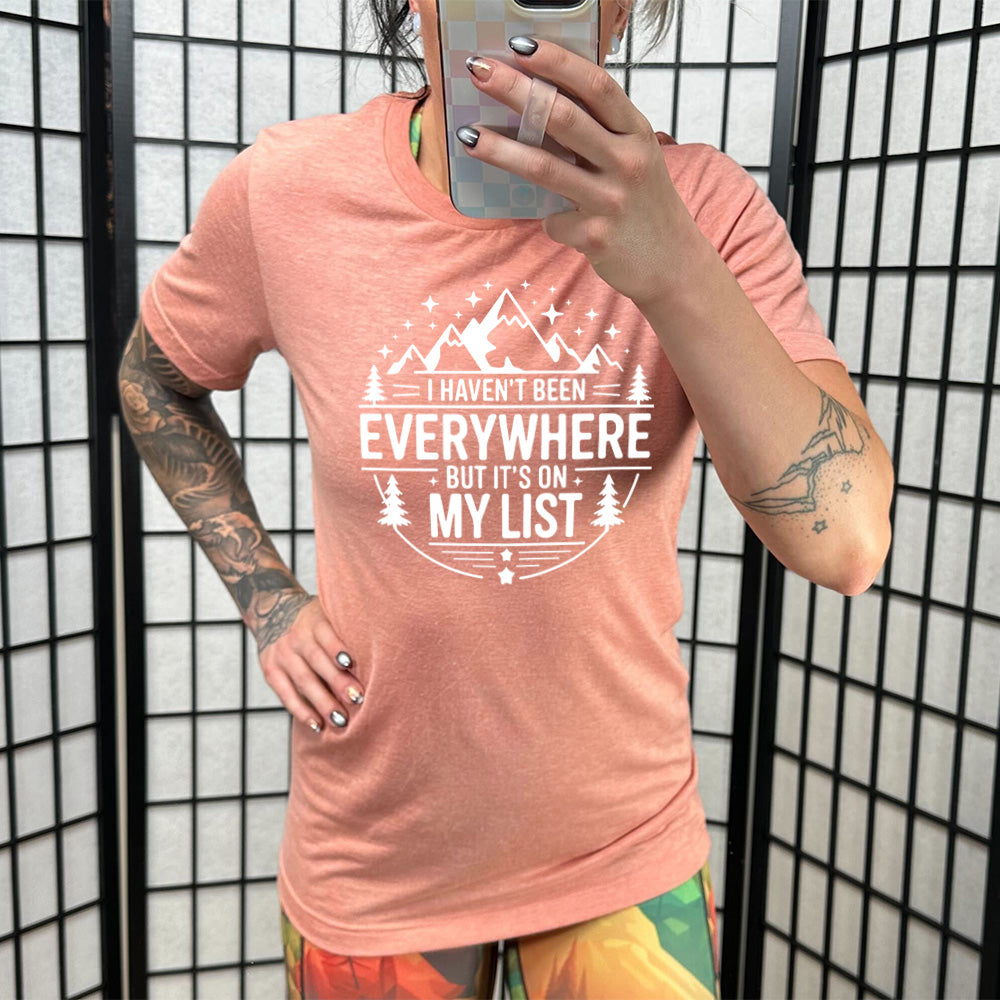 sunset unisex tee with the words "I haven't been everywhere but it's on my list" as part of a circular design involving the graphic of small mountains on top and a few trees on the sides and stars scattered around