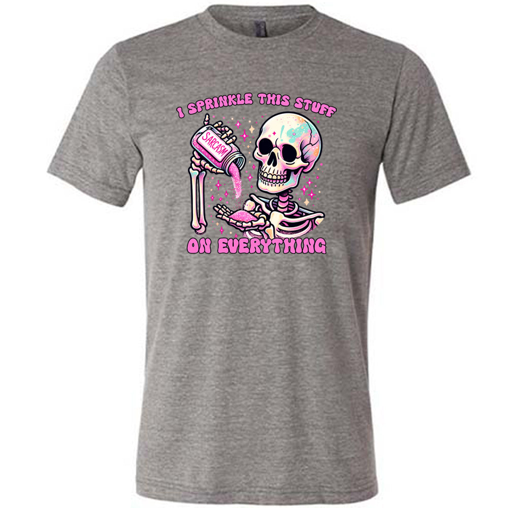 grey unisex tee with the words "I sprinkle this stuff on everything" on it, and the graphic of a skeleton who is pouring a jar labeled "sarcasm" filled with pink glittery material into its hand, surrounded by little pink and white stars