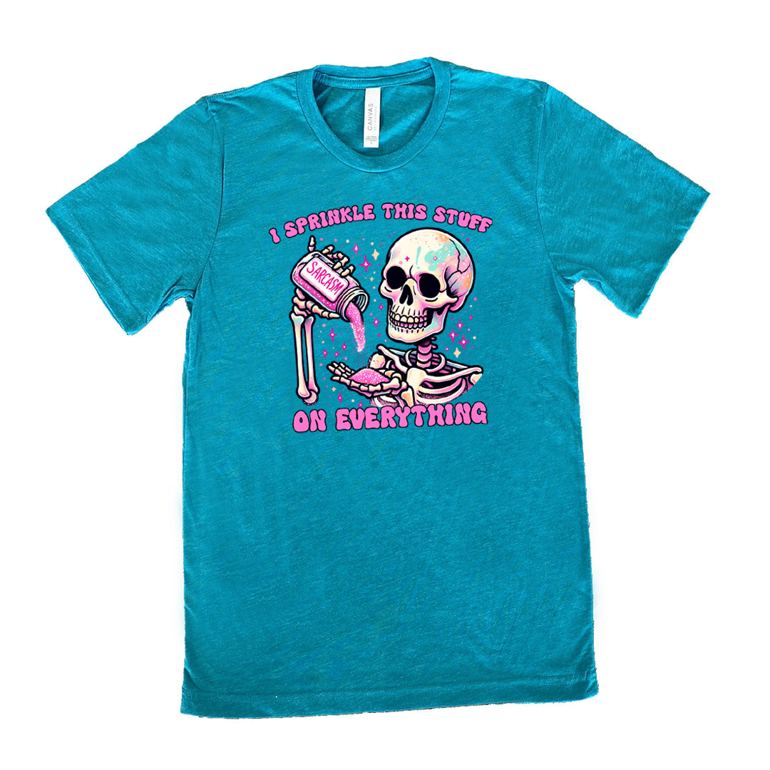 teal unisex tee with the words "I sprinkle this stuff on everything" on it, and the graphic of a skeleton who is pouring a jar labeled "sarcasm" filled with pink glittery material into its hand, surrounded by little pink and white stars