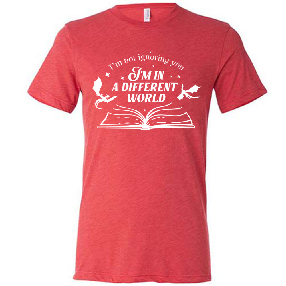 red unisex tee with the words "I'm not ignoring you I'm in a different world" on it, with the graphic of a large book, lying open, and two little dragons flying next to the words, a few stars scattered around with the words "I'm not ignoring you I'm in a different world" on it, with the graphic of a large book, lying open, and two little dragons flying next to the words, a few stars scattered around
