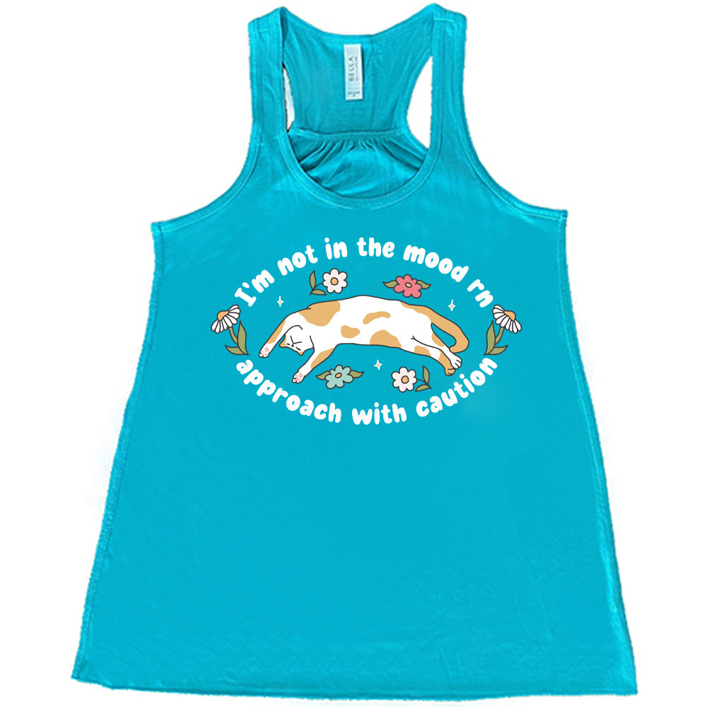 teal racerback tank with the words "I'm not in the mood rn approach with caution" on it in an oval, with the graphic of a cat splayed on its back within the words, and a few flowers around