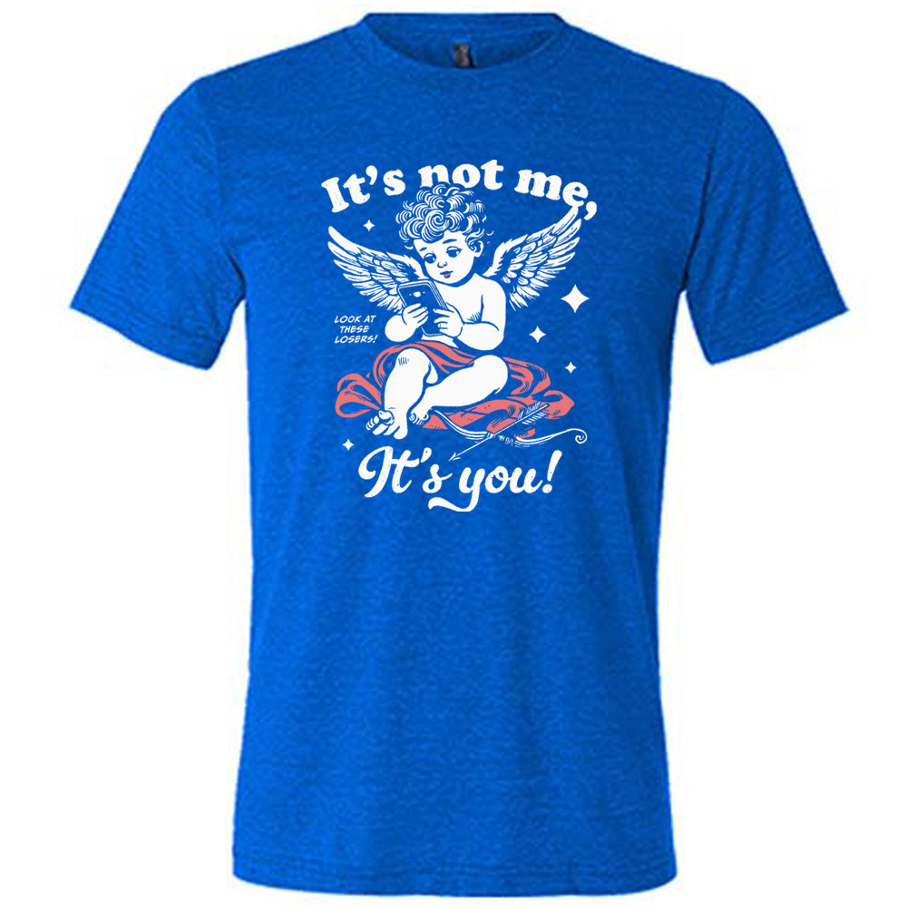 blue unisex tee with the words "it's not me, it's you!" on it, with graphics of Cupid looking at a cell phone (presumably looking at dating apps), with small words saying "look at these losers!" and Cupid's bow and arrow at his side