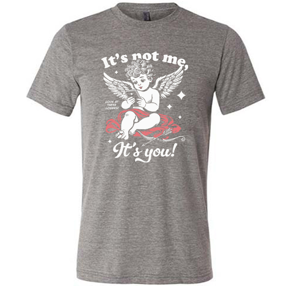 grey unisex tee with the words "it's not me, it's you!" on it, with graphics of Cupid looking at a cell phone (presumably looking at dating apps), with small words saying "look at these losers!" and Cupid's bow and arrow at his side