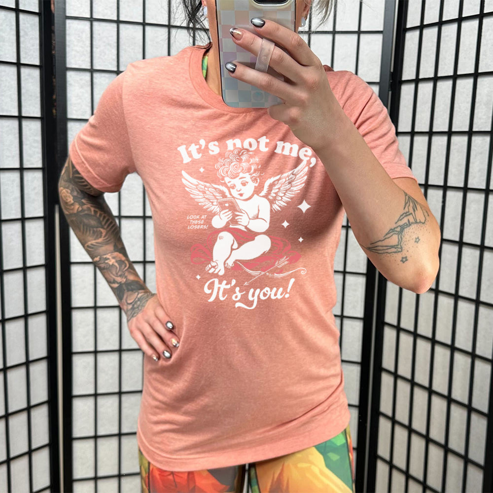 sunset unisex tee with the words "it's not me, it's you!" on it, with graphics of Cupid looking at a cell phone (presumably looking at dating apps), with small words saying "look at these losers!" and Cupid's bow and arrow at his side