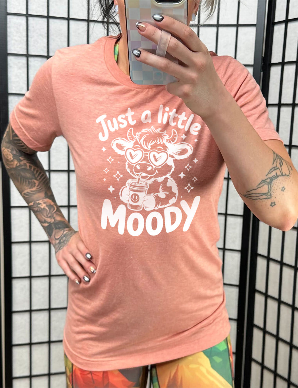 sunset unisex tee with the graphic of a cartoon cow, wearing heart sunglasses and drinking out of an iced cup with a straw, surrounded by little stars, as well as the words "just a little moody" around it