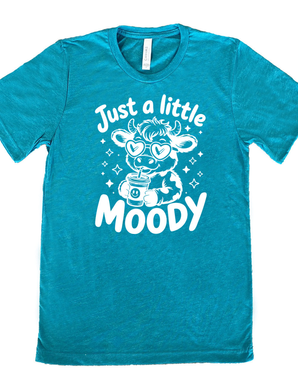teal unisex tee with the graphic of a cartoon cow, wearing heart sunglasses and drinking out of an iced cup with a straw, surrounded by little stars, as well as the words "just a little moody" around it