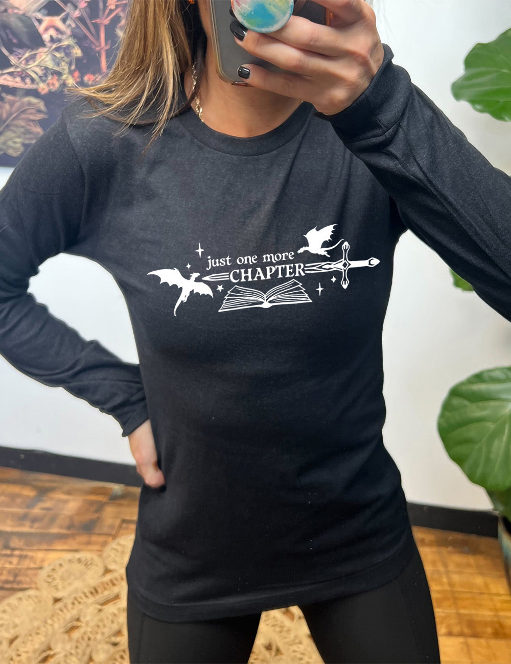 heather black long sleeve shirt with the graphic of an open book, with stars and a couple dragon silhouettes, and a sword, and the words "just one more chapter" on it, with the word "chapter" overlaying the sword