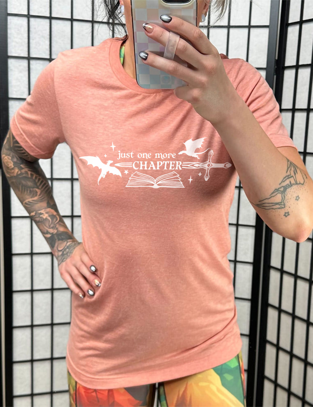 sunset unisex tee with the graphic of an open book, with stars and a couple dragon silhouettes, and a sword, and the words "just one more chapter" on it, with the word "chapter" overlaying the sword