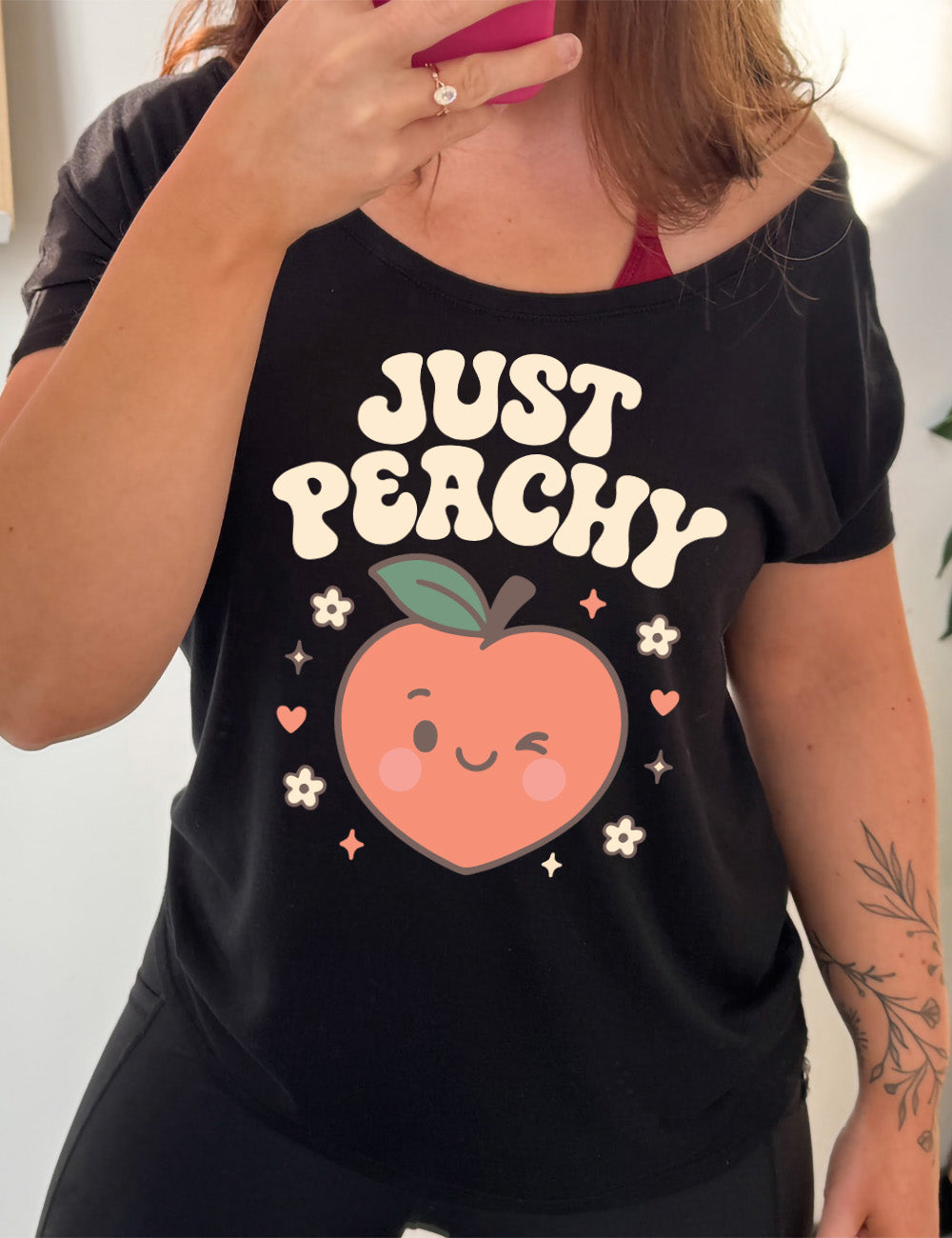 black slouchy tee with the graphic of a winking peach, surrounded by little hearts, flowers, and stars, with the words above it saying "just peachy"