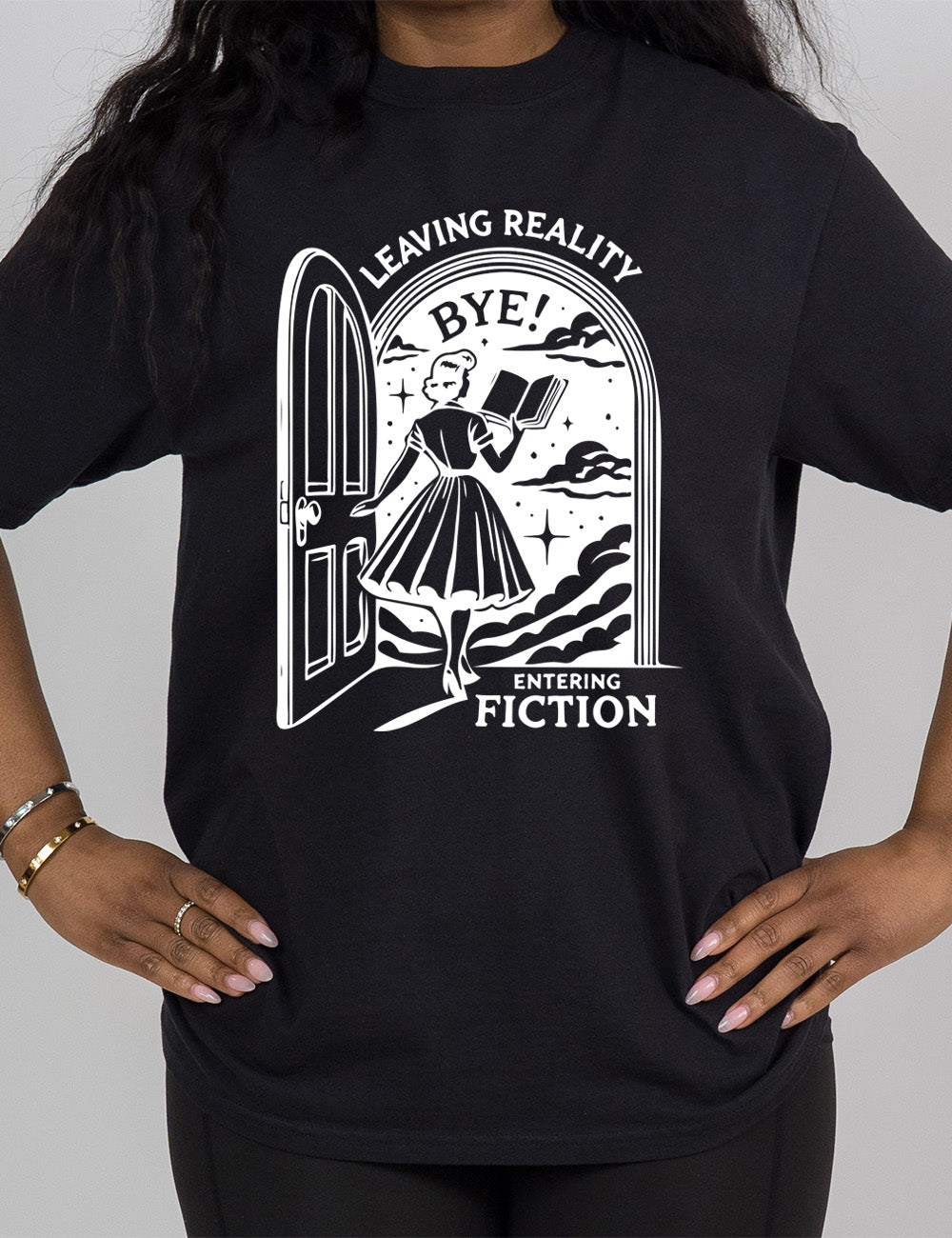 black modern tee with the graphic of a woman, reading a book, and walking through an open door into a fantasy world, with the words "leaving reality entering fiction" on it, and the word "bye!" above the woman's head
