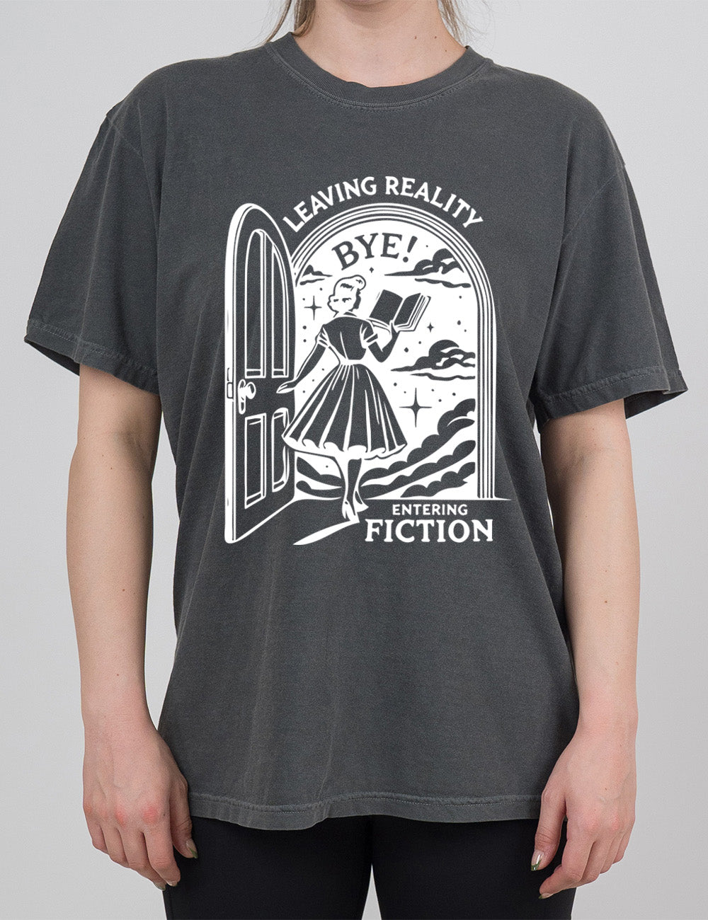 pepper modern tee with the graphic of a woman, reading a book, and walking through an open door into a fantasy world, with the words "leaving reality entering fiction" on it, and the word "bye!" above the woman's head