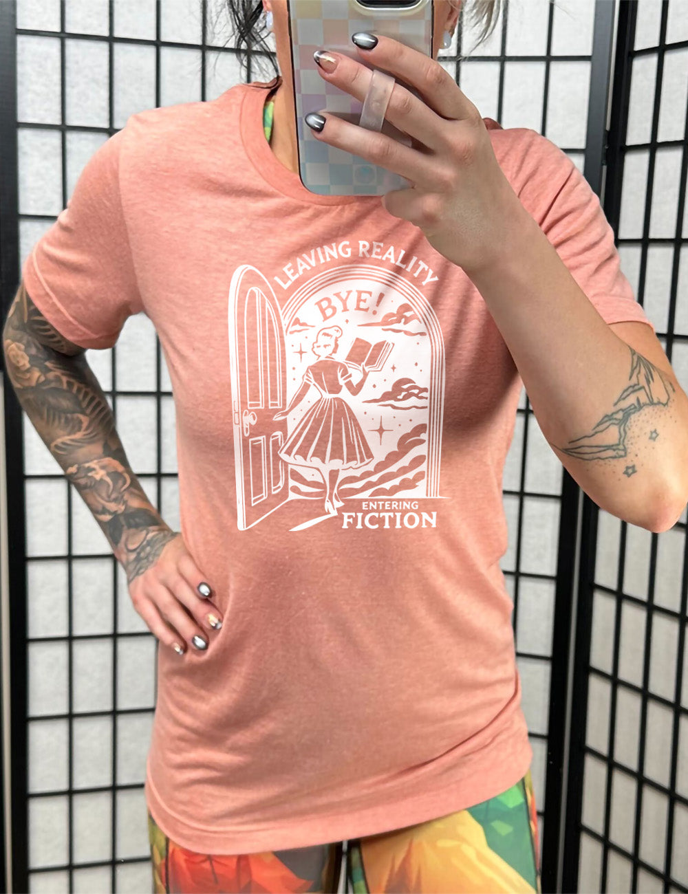 sunset unisex tee with the graphic of a woman, reading a book, and walking through an open door into a fantasy world, with the words "leaving reality entering fiction" on it, and the word "bye!" above the woman's head