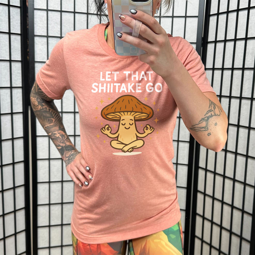 sunset unisex tee with the words "let that shiitake go" on it and the graphic of a mushroom doing a yoga pose, surrounded by little stars