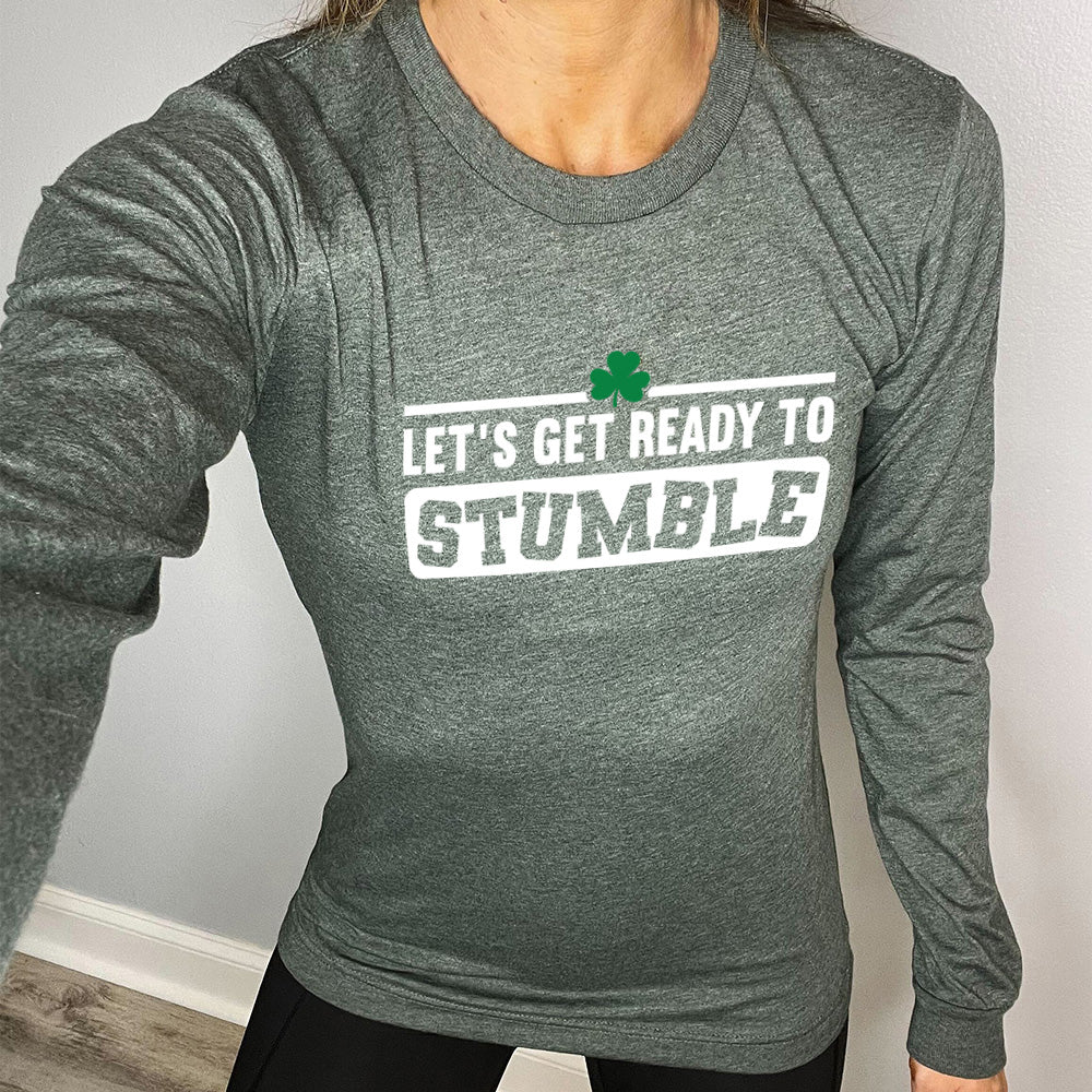 heather forest long sleeve with the words "let's get ready to stumble" on it, with one little shamrock graphic, and the word "stumble" in a rectangular block, and the letters leaning in different directions