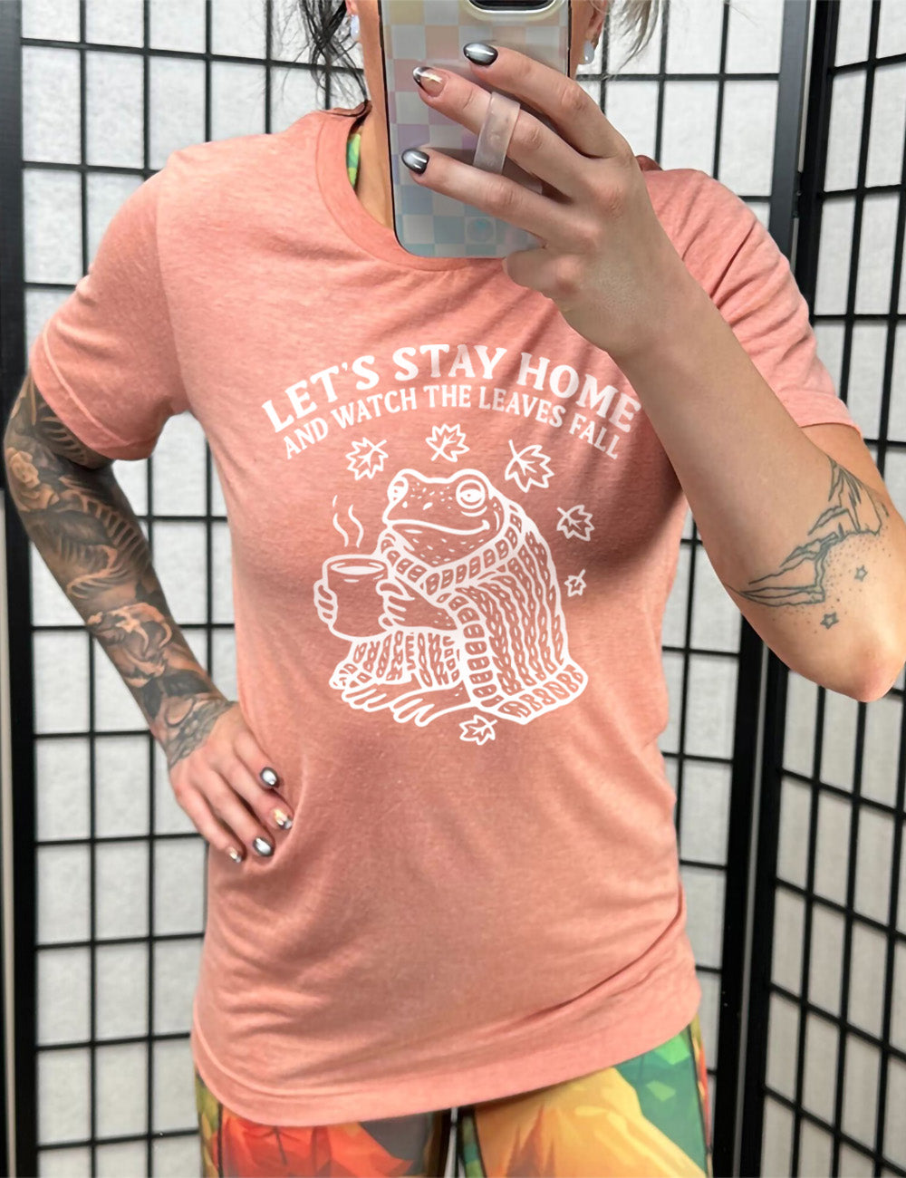 sunset unisex tee with the words "let's stay home and watch the leaves fall" on it, and the graphic underneath of a frog, wrapped in a blanket, holding a mug of steaming beverage, with five leaves arching over it