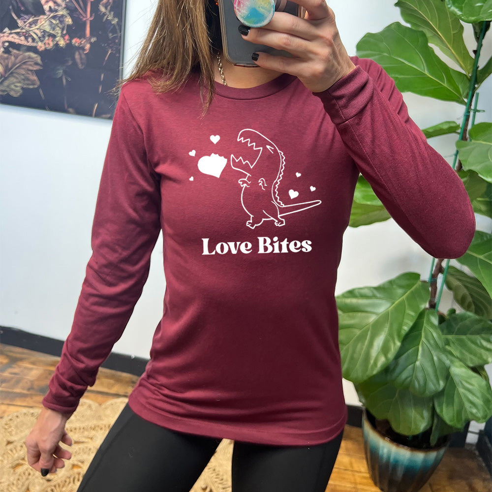 heather cardinal long sleeve shirt with the graphic of a dinosaur outline, eyes shut, mouth wide open, having bitten a chunk out of a heart, with a few other little hearts around it, and the text underneath saying "love bites"