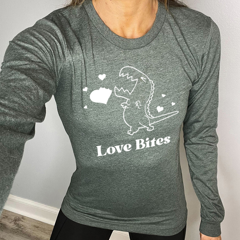 heather forest long sleeve shirt with the graphic of a dinosaur outline, eyes shut, mouth wide open, having bitten a chunk out of a heart, with a few other little hearts around it, and the text underneath saying "love bites"