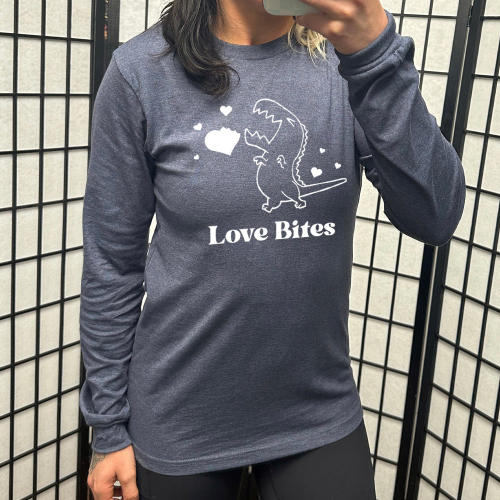 heather navy long sleeve shirt with the graphic of a dinosaur outline, eyes shut, mouth wide open, having bitten a chunk out of a heart, with a few other little hearts around it, and the text underneath saying "love bites"