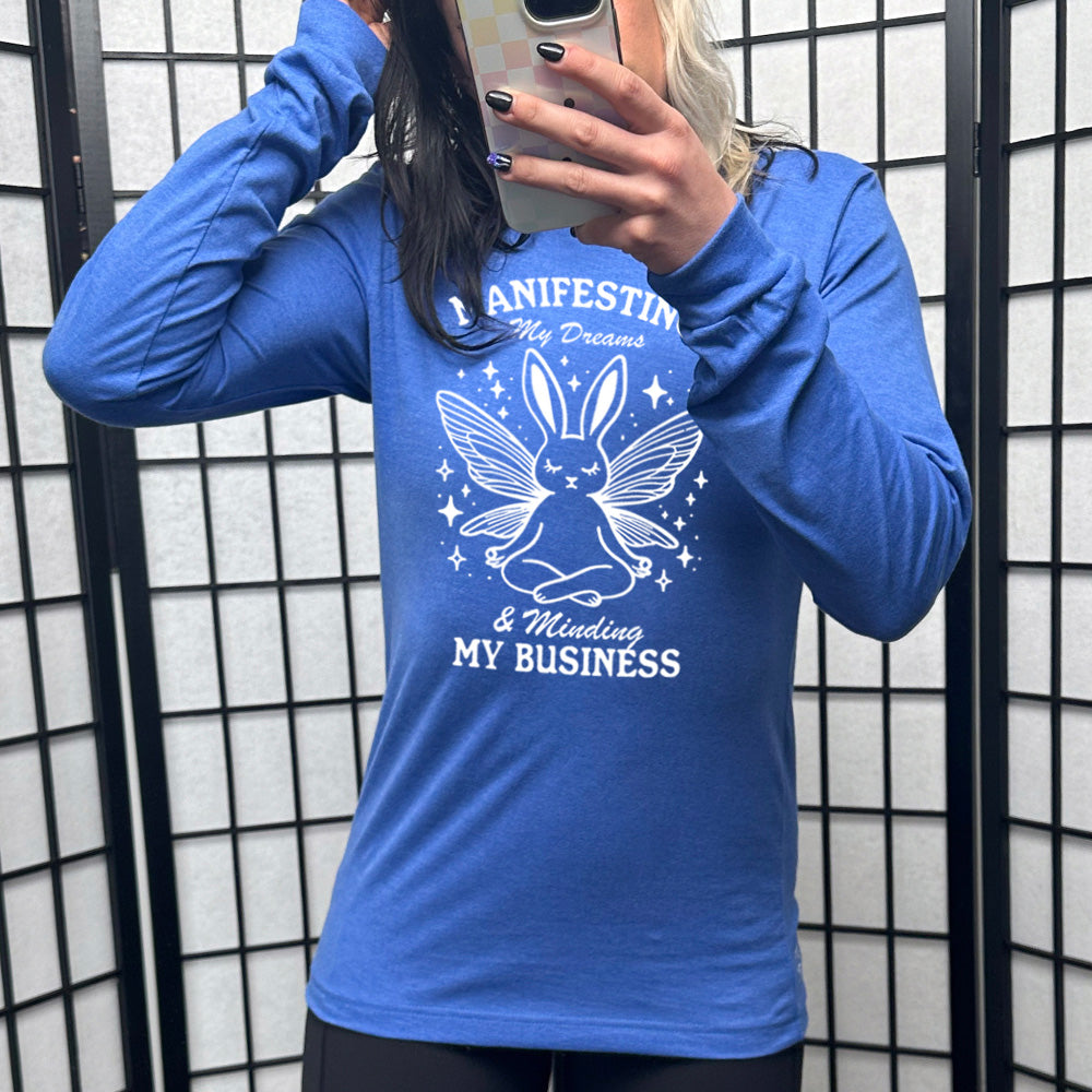 Heather true royal long sleeve shirt with the words "Manifesting My Dreams & Minding My Business" and the graphic of a meditating rabbit with butterfly wings