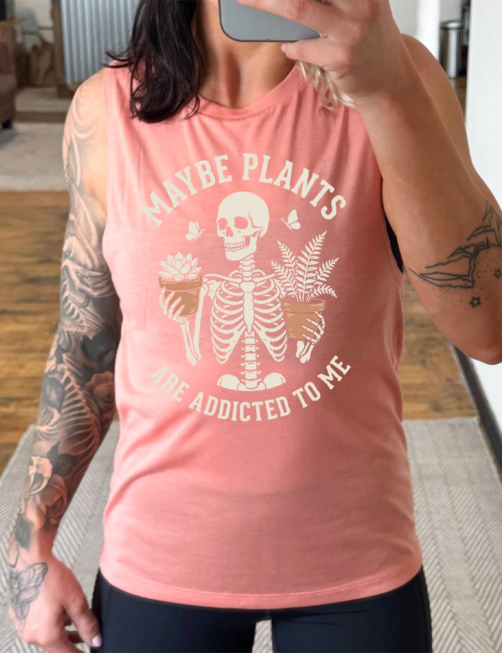 peach muscle tank with the graphic of a skeleton, torso up, holding a houseplant in each hand, with the words around this saying "maybe plants are addicted to me"