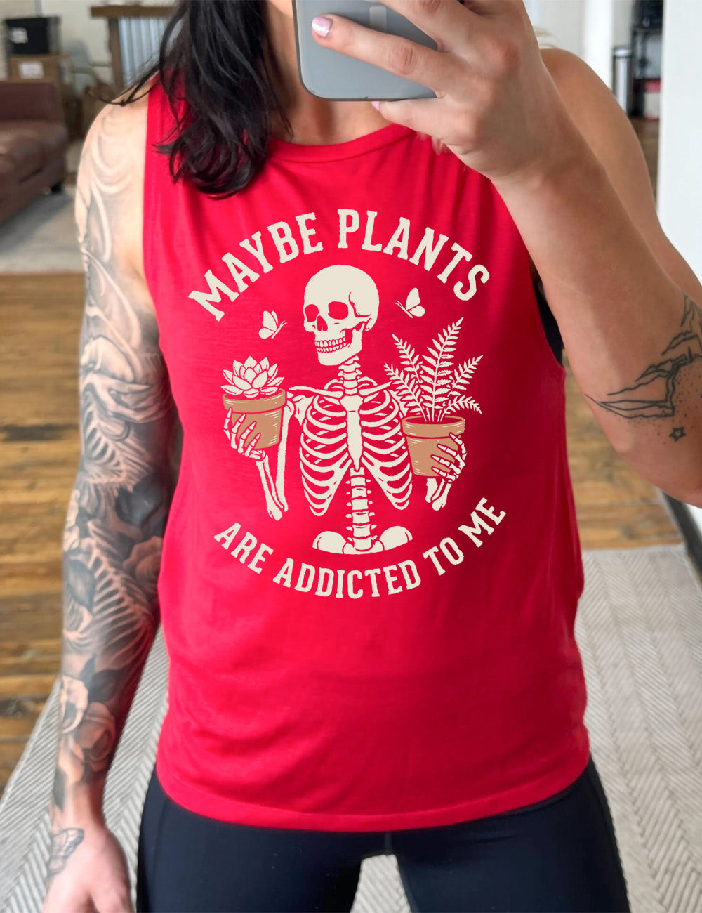 red muscle tank with the graphic of a skeleton, torso up, holding a houseplant in each hand, with the words around this saying "maybe plants are addicted to me"