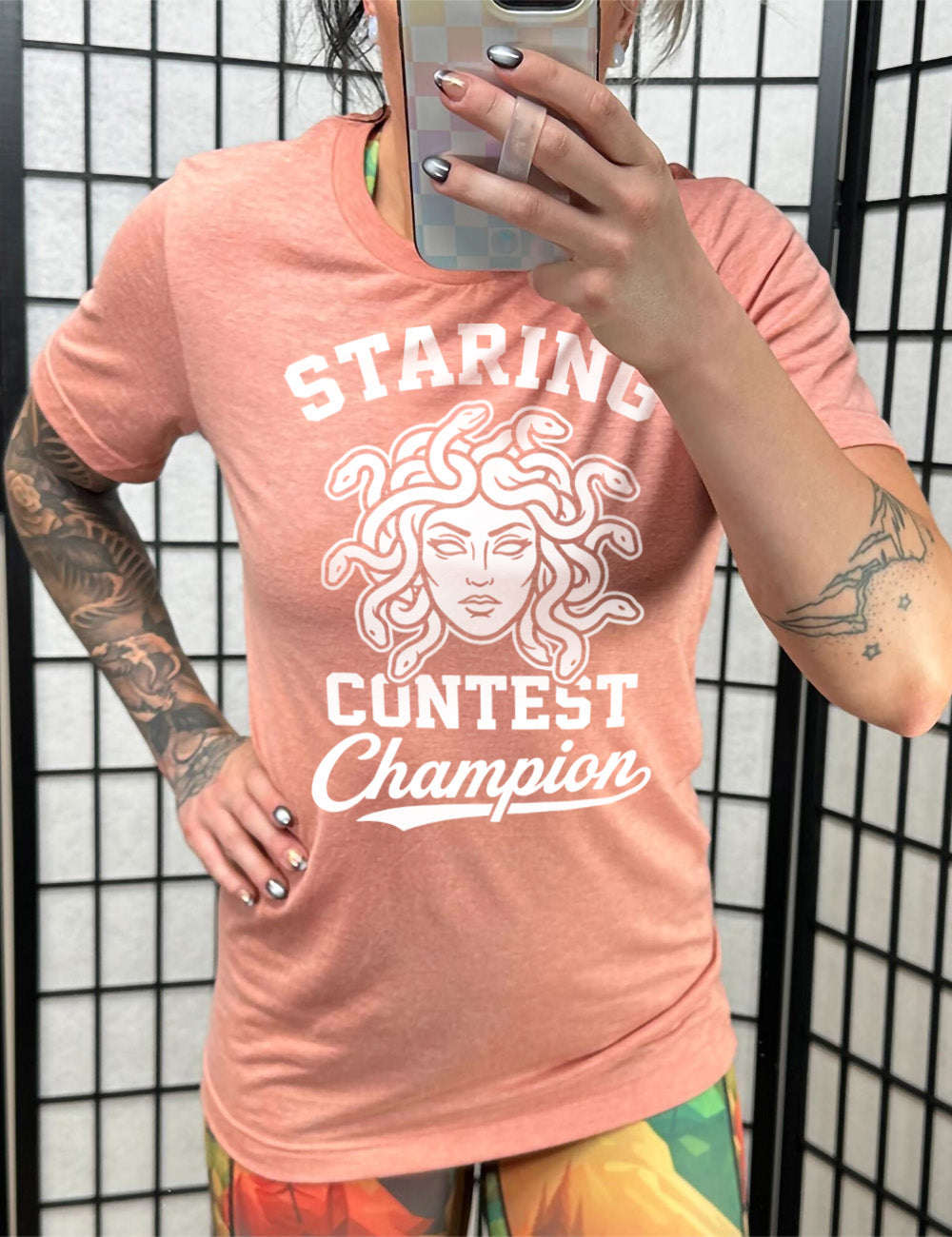 sunset unisex tee with words surrounding the graphic of medusa's head saying "staring contest champion"