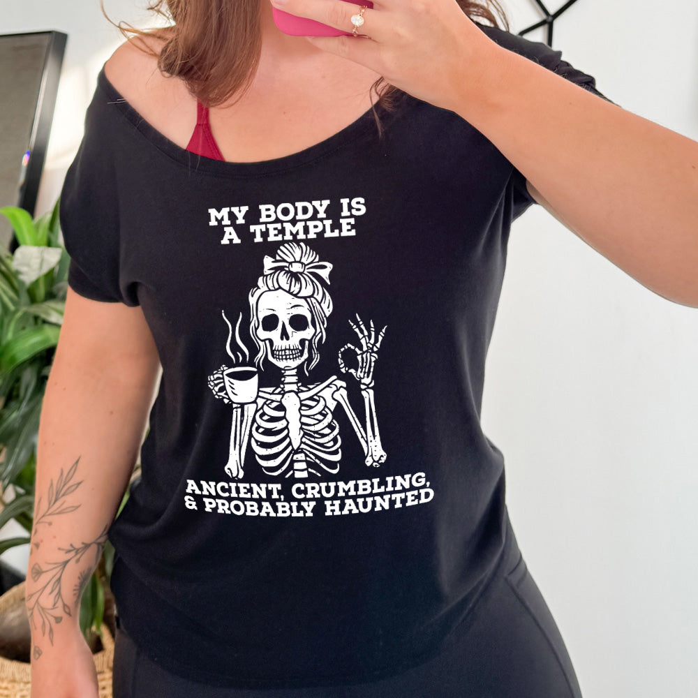 black slouchy tee with the words "My Body Is a Temple Ancient, Crumbling, & Probably Haunted" on it, with the graphic of a skeleton with its hair in a bow, with one hand holding steaming coffee, and the other giving the OK symbol