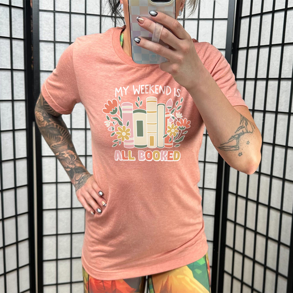 Model wearing sunset colored shirt with books and flowers graphic on it that says "My Weekend Is All Booked"