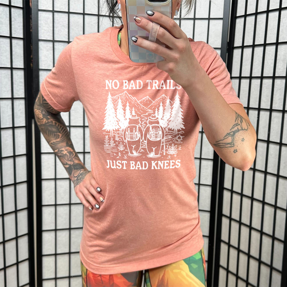 Model wearing sunset colored shirt with two bears hiking graphic on it that says "No Bad Trails Just Bad Knees"
