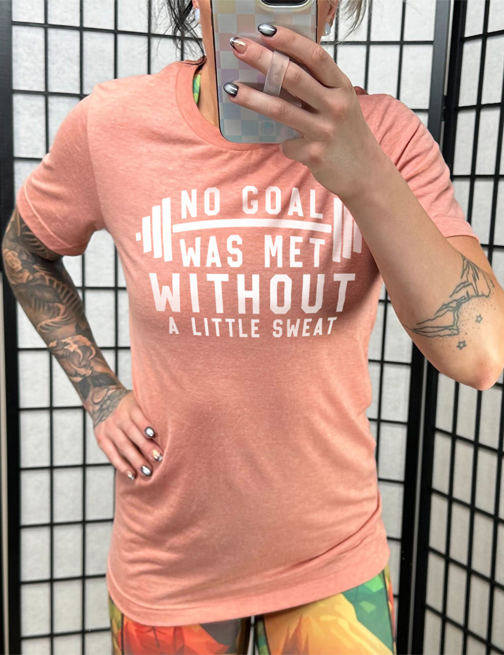 sunset unisex tee with the words "NO GOAL WAS MET WITHOUT A LITTLE SWEAT" on it, with a barbell graphic after the words "no goal"