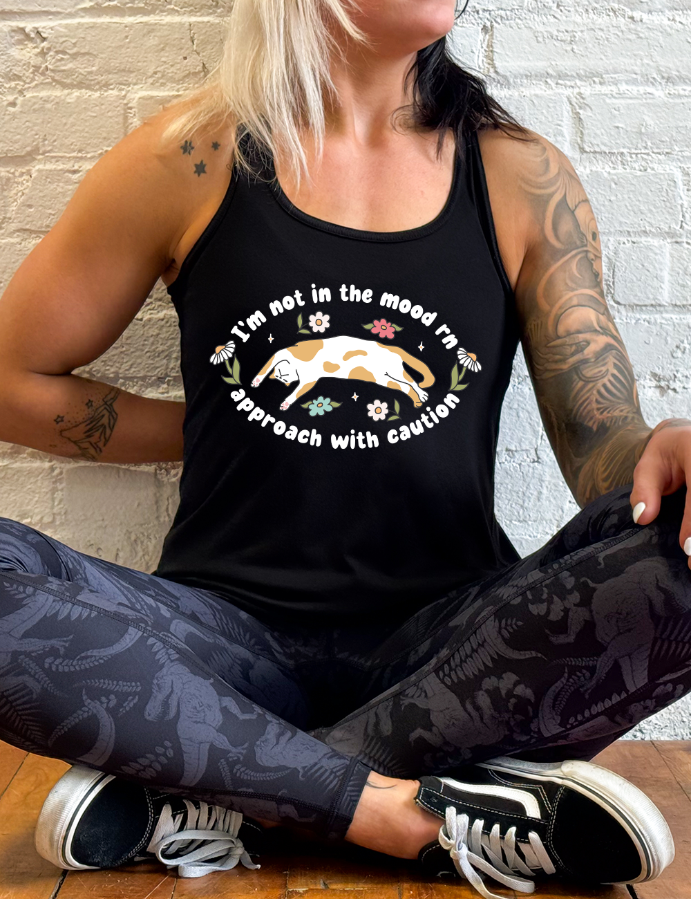 black racerback tank with the words "I'm not in the mood rn approach with caution" on it in an oval, with the graphic of a cat splayed on its back within the words, and a few flowers around