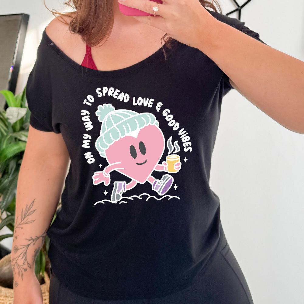 black slouchy shirt with the words "on my way to spread love & good vibes" on it, with graphics of a cheerful, cartoony heart strolling along with a mug of a hot drink steaming in its hand, a hat on its head, and shoes on