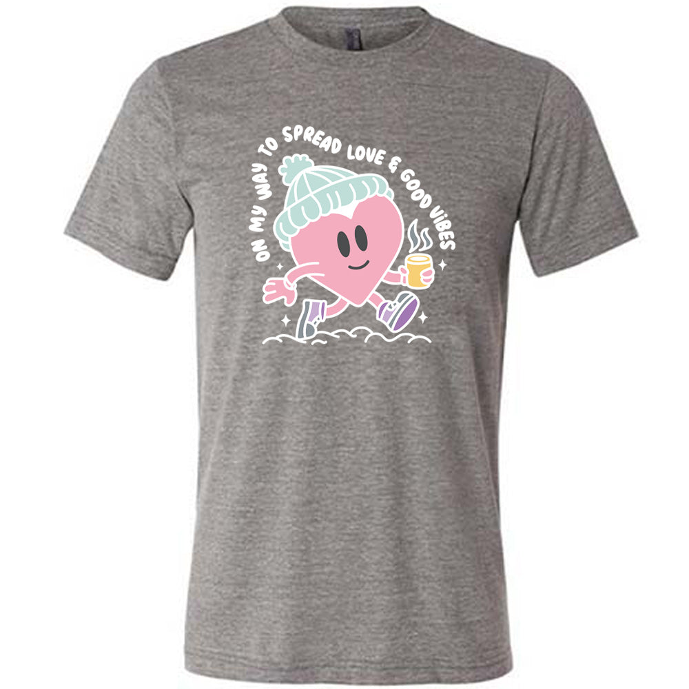 grey unisex tee with the words "on my way to spread love & good vibes" on it, with graphics of a cheerful, cartoony heart strolling along with a mug of a hot drink steaming in its hand, a hat on its head, and shoes on