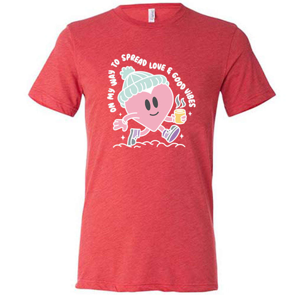 red unisex tee with the words "on my way to spread love & good vibes" on it, with graphics of a cheerful, cartoony heart strolling along with a mug of a hot drink steaming in its hand, a hat on its head, and shoes on
